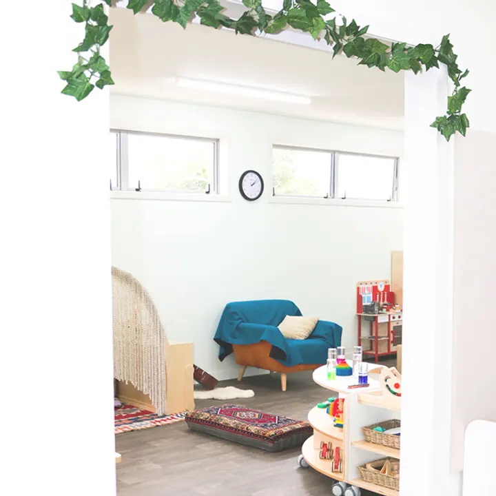 A quiet corner seen through a doorway trailed with ivy: a small blue sofa, a woven canopy, rugs on the floor and a clock on the wall.