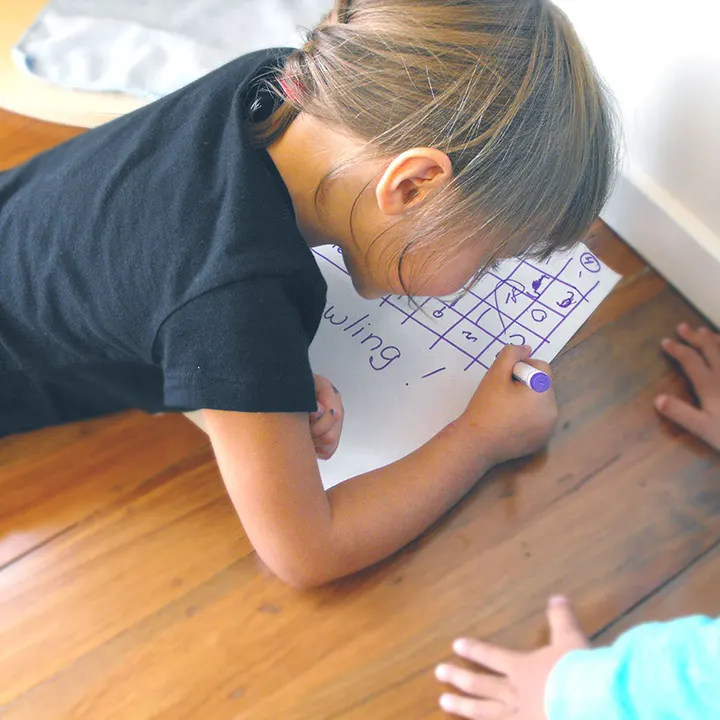 A child lying on a wooden floor, drawing a grid of letters on a large sheet of paper with a marker, with another child’s hands at the edge of the picture.